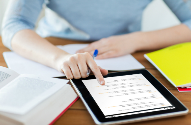A woman sits at a table holding a pen and scrolling on a tablet. An open book and notepad are also on the table.