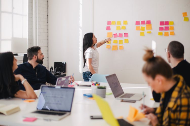 A group of people sit around a table with open laptops and take notes on sticky notes. They look to a women who is standing in front of them and gesturing towards multiple sticky notes stuck to a wall.
