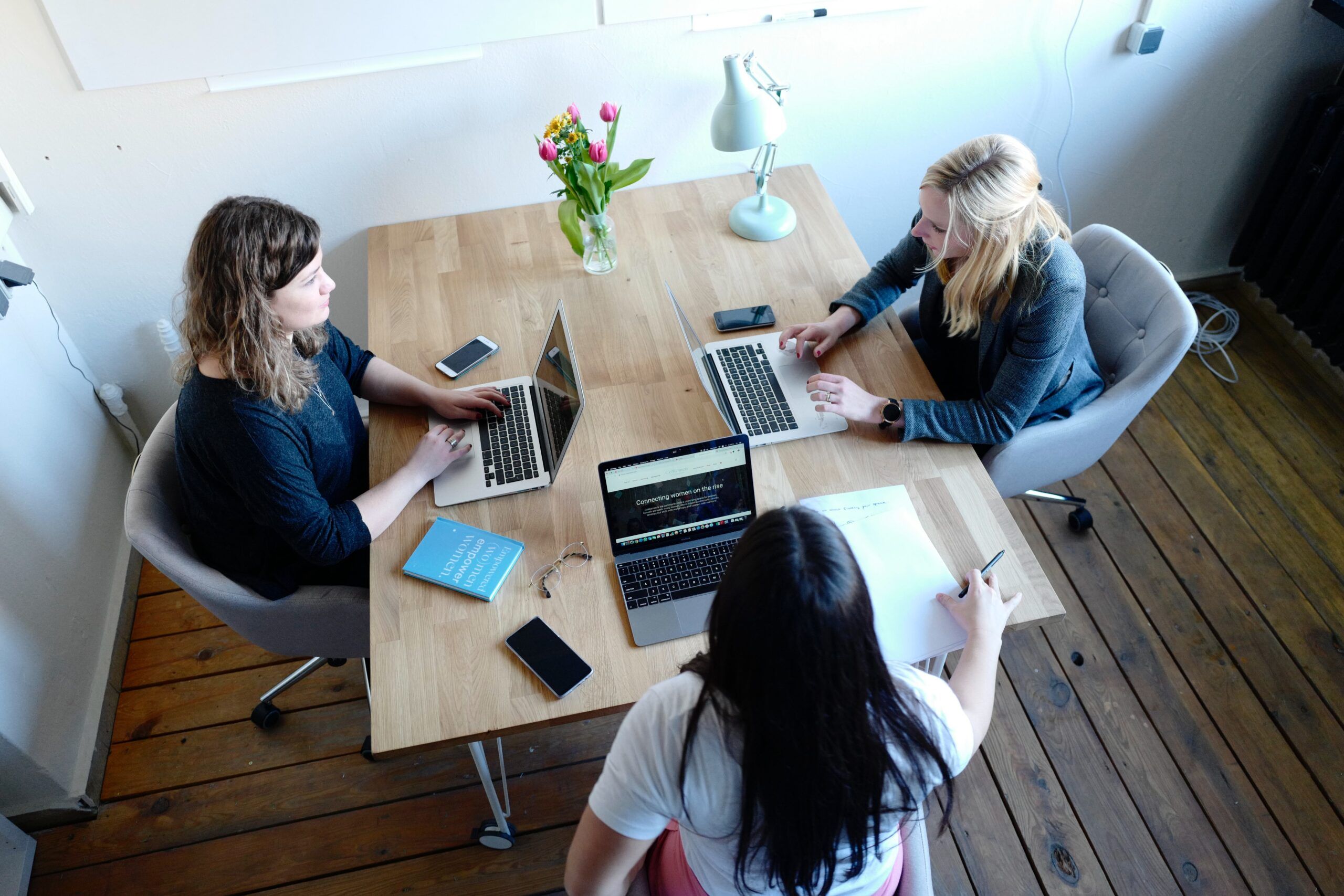 Three women sitting at a table with an open laptop in front of each of them. One woman holds a pen above a sheet of paper.