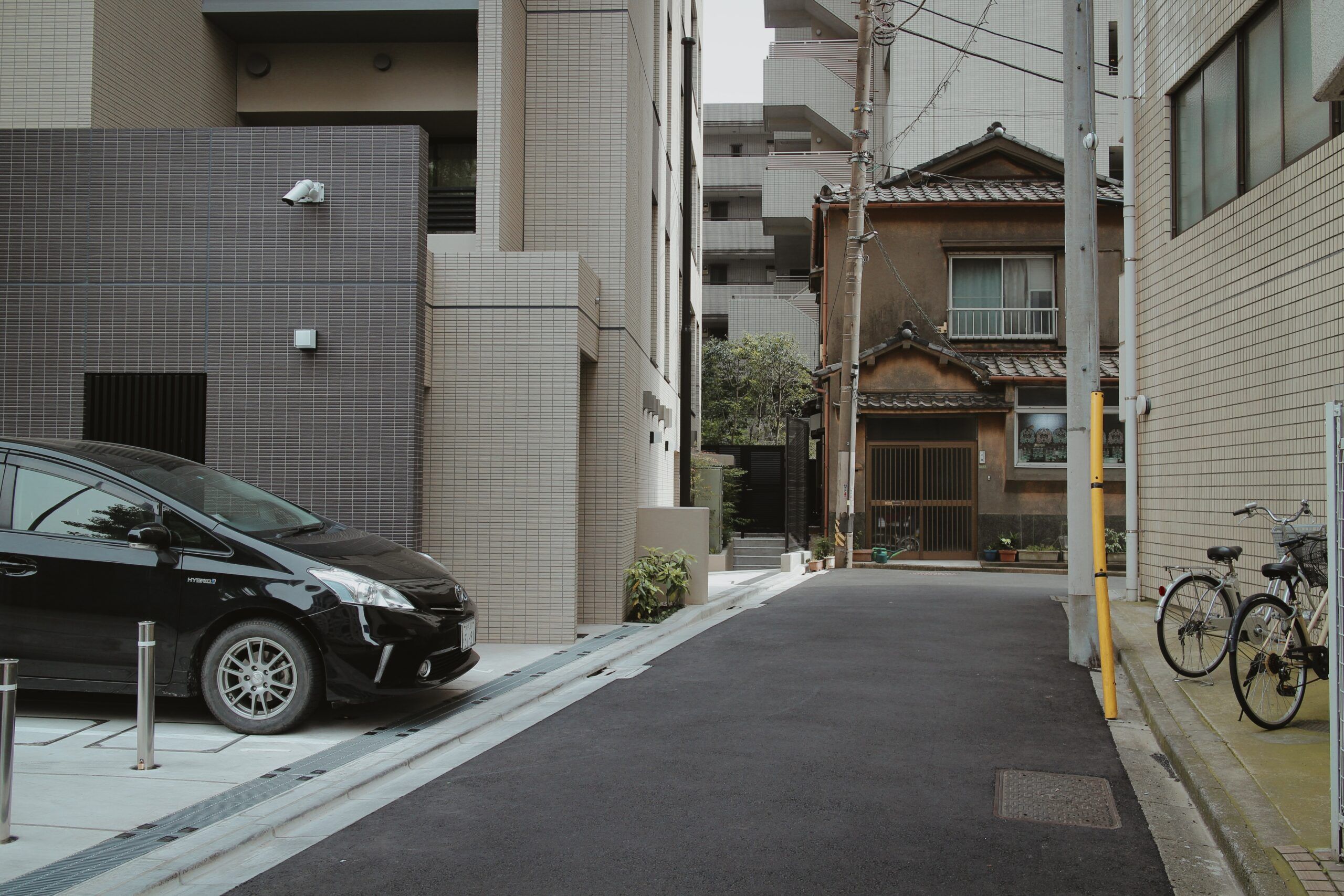 A narrow street in a city. On the left side of the street is a building with a car parked next to it. On the right side of the street is a building with two bicycles parked in front of it. At the end of the street is a house.